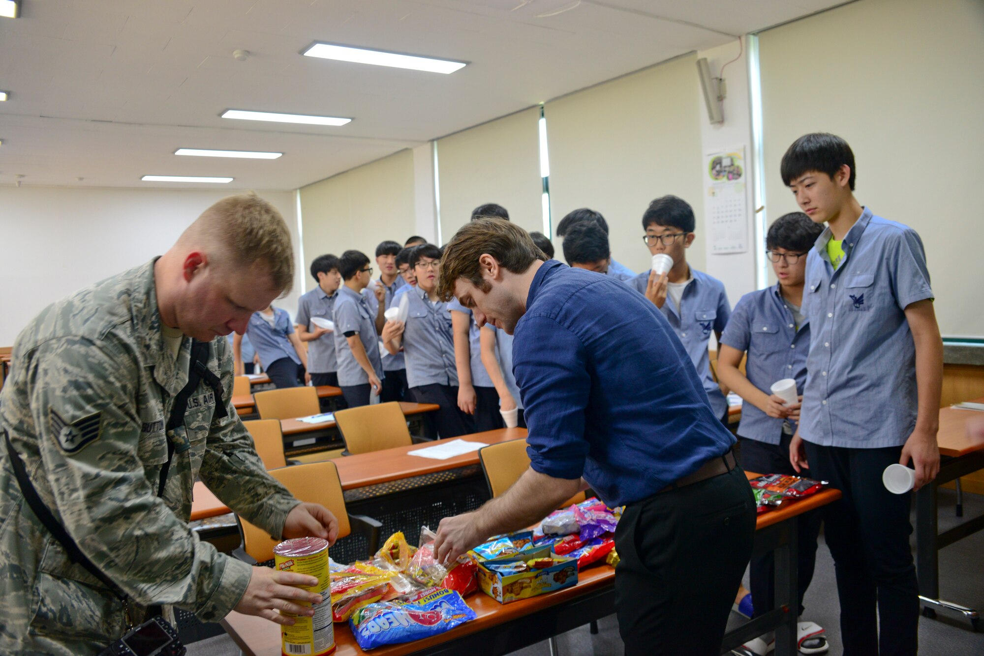 Staff Sgt. Benjamin Sutton, 51st Fighter Wing public affairs photojournalist, helps Chris Binnie, English as a foreign language teacher at Cheonan High School, Cheonan, South Korea, set up a table of July 10, 2015. The students of the podcast club, “Channel K Radio," tried various American snacks during an international cultural exchange between Cheonan and Warren Academy, McMinnville, Tenn. The exchange allowed the Korean students to not only try American snacks, but to meet Airmen on a friendly and personal level in understanding the mission of the U.S. Air Force in Korea. (U.S. Air Force photo/Senior Airman Kristin High)