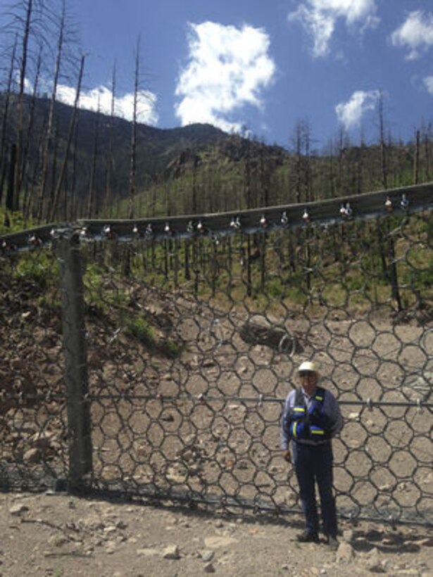 As Climate Changes, Santa Clara Strives To Become Resilient To Worsening Fires, Floods: Santa Clara Pueblo Gov. Michael Chavarria stands before one of four debris collection structures installed this past winter to help protect infrastructure and roads downstream. The structure captures sediment, boulders and trees washed down the mountain during flash floods. New flood scars carved into the peaks above weren’t easily visible at first, but lateral movement of the water has carved them into wide ravines. ‘Now,’ says Chavarria, ‘you can see them from the community.’ Margaret Wright/The New Mexican