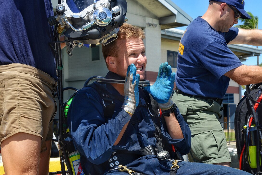 Navy Petty Officer 1st Class David Partin, seated, receives his diving helmet that will attach to the collar he's already wearing before entry into the waters of Honolulu Harbor from the pier at Coast Guard Base Honolulu, July 30, 2015.