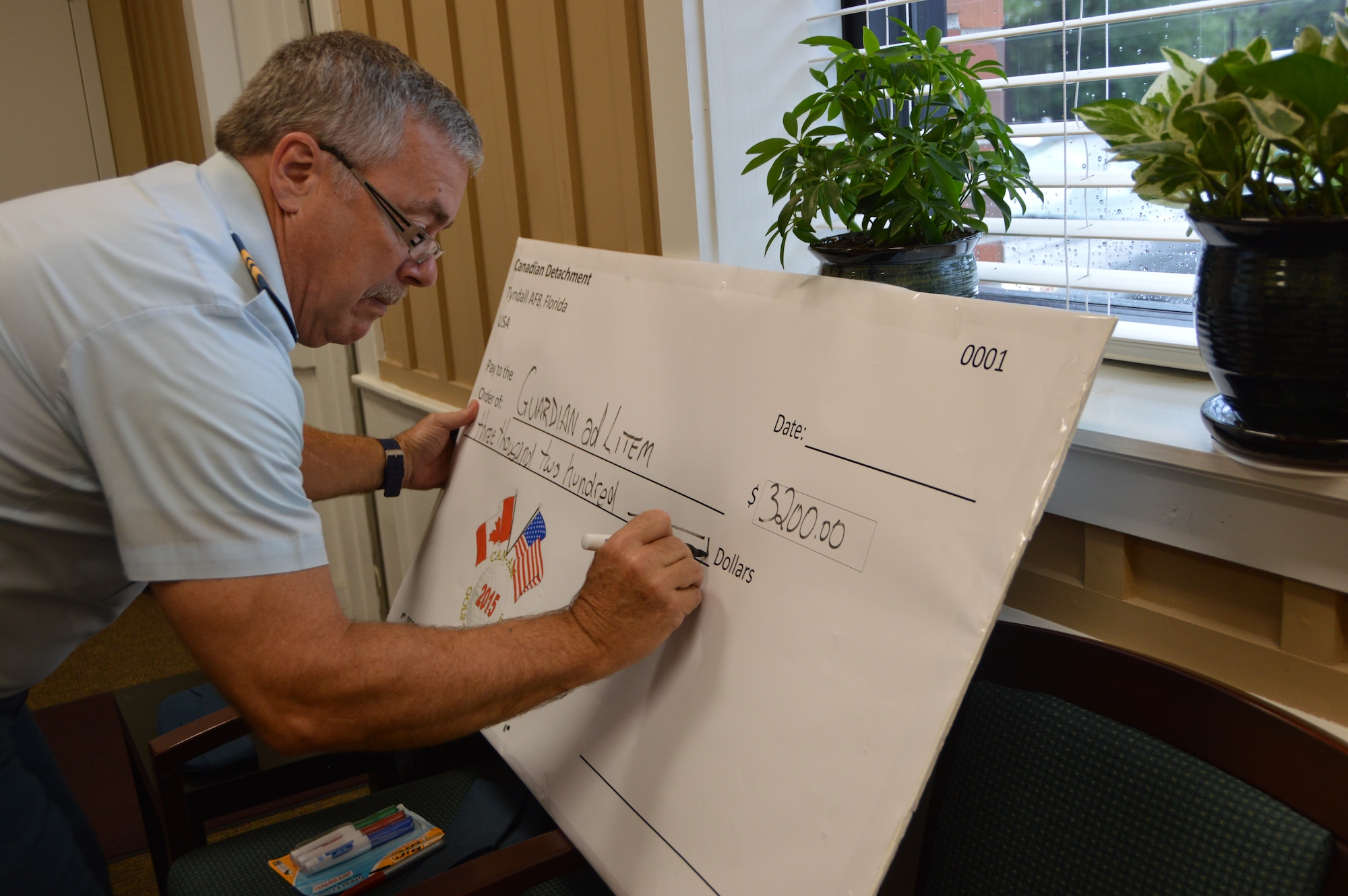Maj. Trent Morrisey, of the Assessments, Lessons Learned and Exercises Directorate, Continental North American Aerospace Defense  Command Region-1st Air Force ( Air Forces Northern), writes the ceremonial check for the donation presentation June 10  to Guardian ad Litem officials. Guardian ad Litem advocates for children in the court system as a result of abuse and neglect.  The donation reflects a portion of the proceeds raised during the  CAN-AM 2015 Golf Tournament May 28.  (Air Force photo released/Photo by Mary McHale)