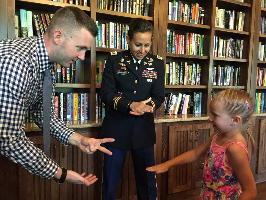 A soldier, a civilian and a girl make hand gestures while standing in front of a bookcase.
