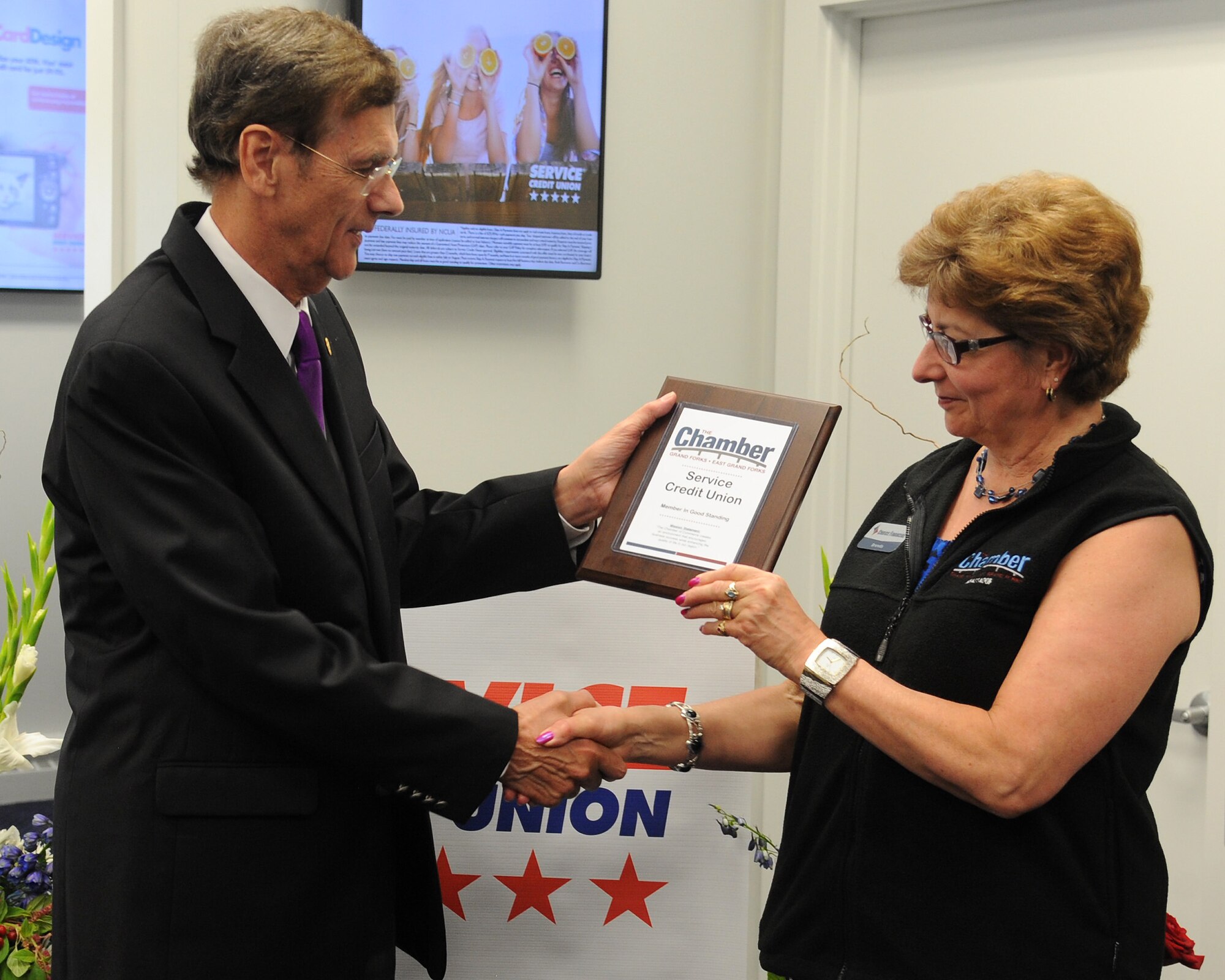 Brenda Kovar, ambassador captain of the Grand Forks welcoming committee, presents Gordon Simmons, president of Service Credit Union, with a “Member in Good Standing” plaque from the Grand Forks and East Grand Forks Chamber of Commerce. Simmons noted that the plaque will be displayed in the SCU museum where numerous other awards and accolades received by the company are also viewable. (U.S. Air Force Photo by Senior Airman Zachiah Roberson/released)