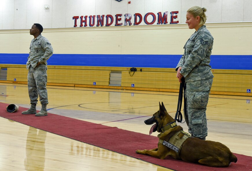 Senior Airman Dominick Young, 799th Security Forces Squadron military working dog handler, Staff Sgt. Anique Brassard, 799 SFS MWD handler, and her partner MWD SSamuel, stand by to give a K9 demonstration as safety rules are given inside the Indian Springs Schools gymnasium March 2, 2014. MWDs and their handlers train together to build a close bond to better protect military personnel and equipment, detect explosives, and conduct search and rescue operations during disasters. (U.S. Air Force photo by Tech. Sgt. Shad Eidson/released)