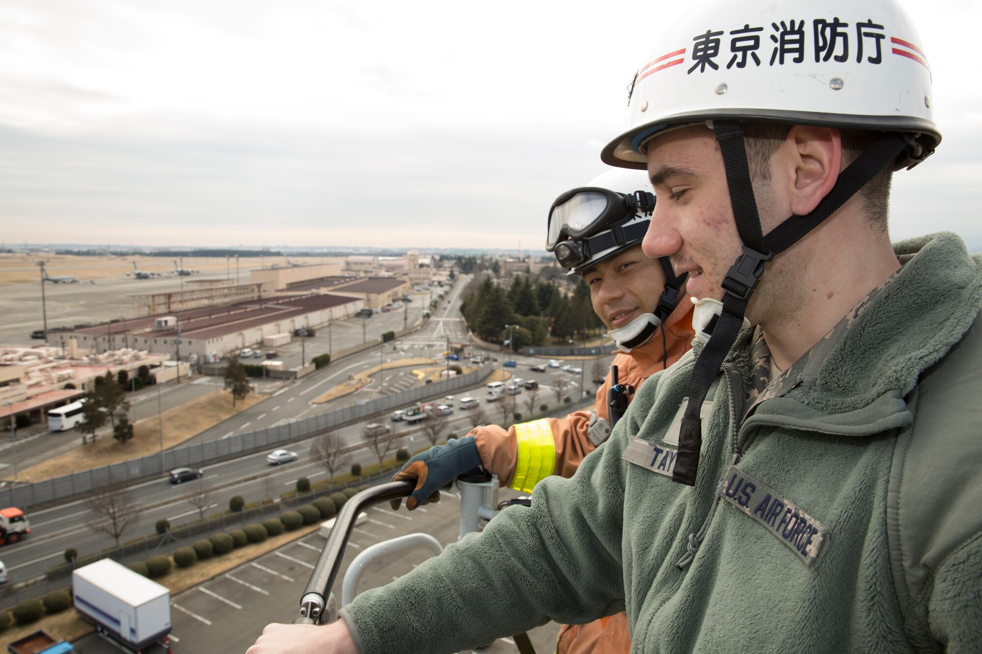 (Left to right) Airman 1st Class Steven Taylor, 374th Civil Engineer Squadron firefighter, and Yuki Yoshizawa, Tokyo Fire Department Fussa Fire Station firefighter, are elevated 100 feet by a Tokyo FD fire ladder truck, during the safety festival at Yokota Air Base, Japan, March 6, 2015. The 374th Airlift Wing Safety Office provided Yokota residents with various events such as the Tokyo Metropolitan Police Department motorcycle unit demonstration, traffic safety demonstration, earthquake and smoke house, firefighting experience, fire ladder trucks experience, 374th Security Forces Squadron patrol vehicle ride, seatbelt simulator and child car seat checks. (U.S. Air Force photo by Osakabe Yasuo/Released) 