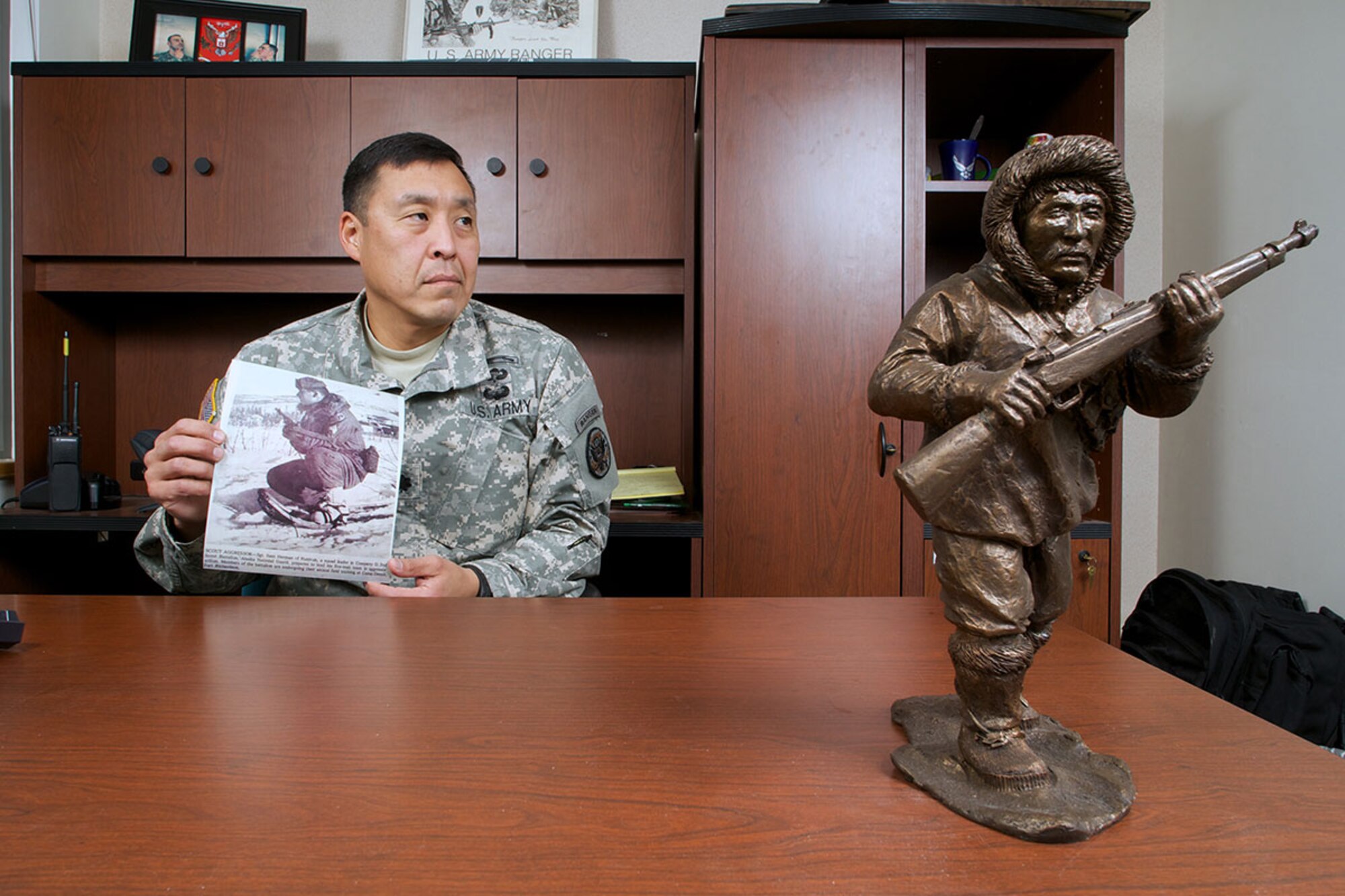 Alaska Army National Guard Lt. Col. Wayne Don, 107th Civil Support Team commander, holds a photo of his uncle, Sam Herman, who served with the Alaska Territorial Guard  during World War II. Herman is pictured during his subsequent service with the Alaska  Army National Guard. A statuette of an ATG scout keeps vigil on Wayne’s desk. (U.S. Air Force photo/David Bedard)