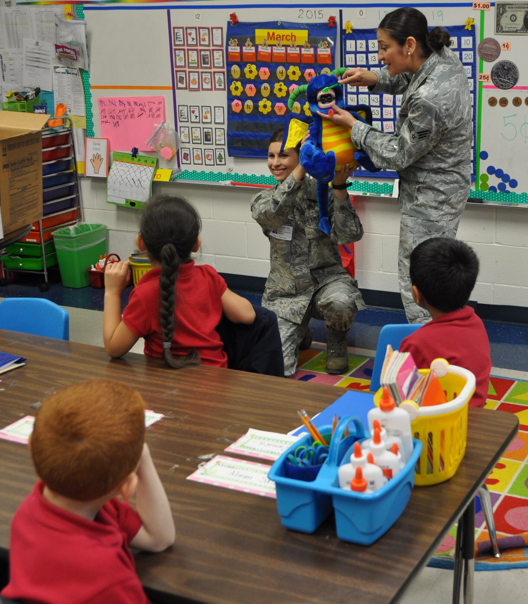 Senior Airmen Stacia Salas (standing) and Tequina Fernandez (kneeling), 433rd Aerospace Medicine Squadron dental assistants, demonstrate to students at Cable Elementary School in San Antonio, Texas March 20, 2015 on how to properly brush their teeth during their presentation. (U.S. Air Force photo by Capt. Cris Medina)