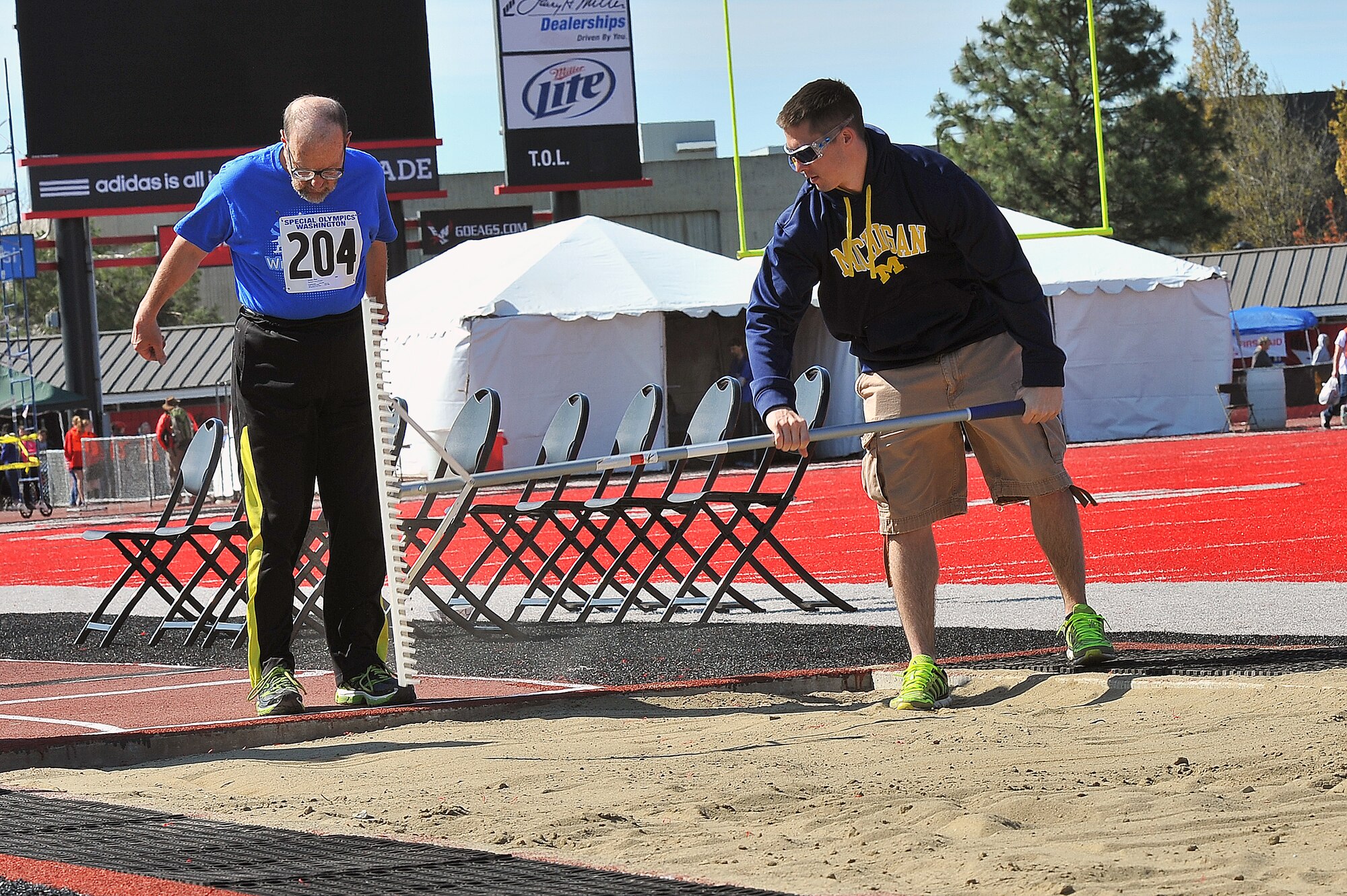 Airman 1st Class Shane Sulier, 92nd Operations Support Squadron airfield systems technician, rakes sand while Don Locke, Spokane Valley Knights participant, prepares to compete in the long jump during the Special Olympics May 2, 2015, in Cheney, Wash. The mission of Special Olympics is to provide year-round sports training and athletic competition in a variety of Olympic-type sports for children and adults with intellectual disabilities, giving them continuing opportunities to develop physical fitness, demonstrate courage, experience joy and participate in a sharing of gifts, skills and friendship with their families, other Special Olympics athletes and the community. (U.S. Air Force photo/Staff Sgt. Veronica Montes)