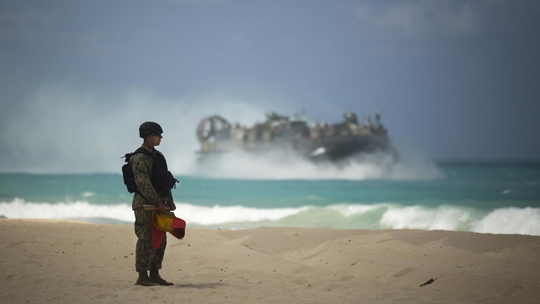 A U.S. Navy service member stands on the beach as a Landing Craft Air Cushion prepares to breach the shore during the MARFORPAC-hosted U.S. Pacific Command Amphibious Leaders Symposium at Marine Corps Training Area Bellows May 19, 2015. The objective of PALS is to have meaningful dialogue on key aspects of maritime and amphibious operations, capability development, and interoperability.