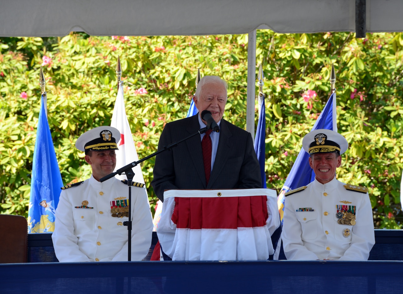 A man in a suit speaks, flanked by sailors.