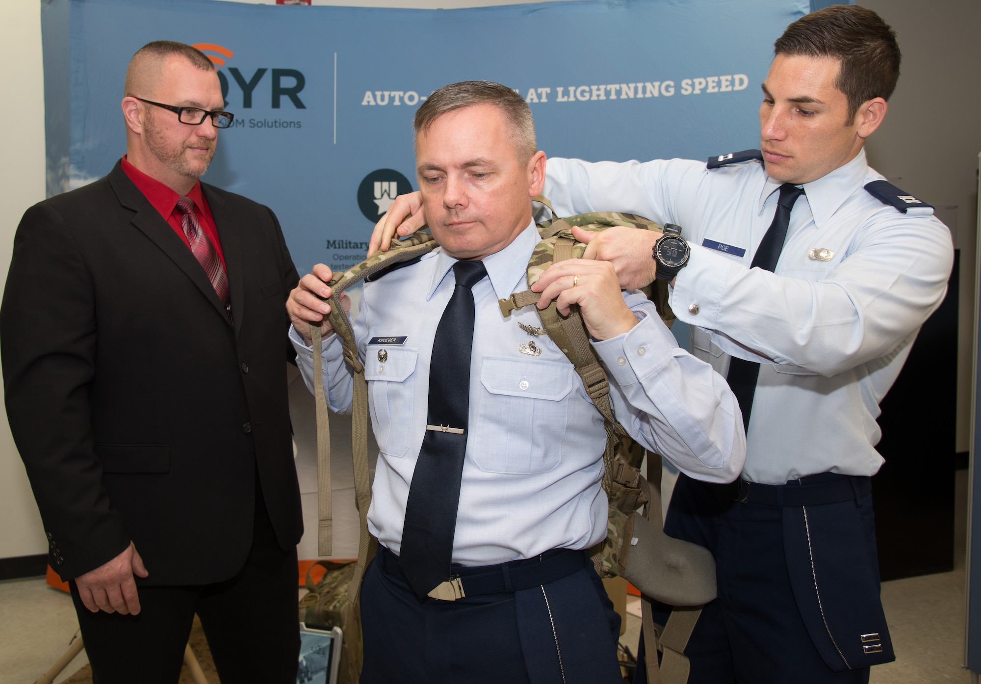 U.S. Air Force Capt. Hayden Poe, far right, a Global Broadcast Service program manager, fastens a GBS rucksack-portable receive suite device onto Col. Todd Krueger, center, Space, Aerial and Nuclear Networks senior materiel leader, during a facility tour of AQYR Technologies in Hollis, N.H., Oct. 30, 2015. AQYR Technologies was recently awarded Hanscom Air Force Base's largest small business innovation research contract to date for the production and delivery of the devices. The receive devices come in a suitcase- and rucksack-portable size that will allow service members to receive near real-time video and data in remote locations. (U.S. Air Force photo by Mark Herlihy)