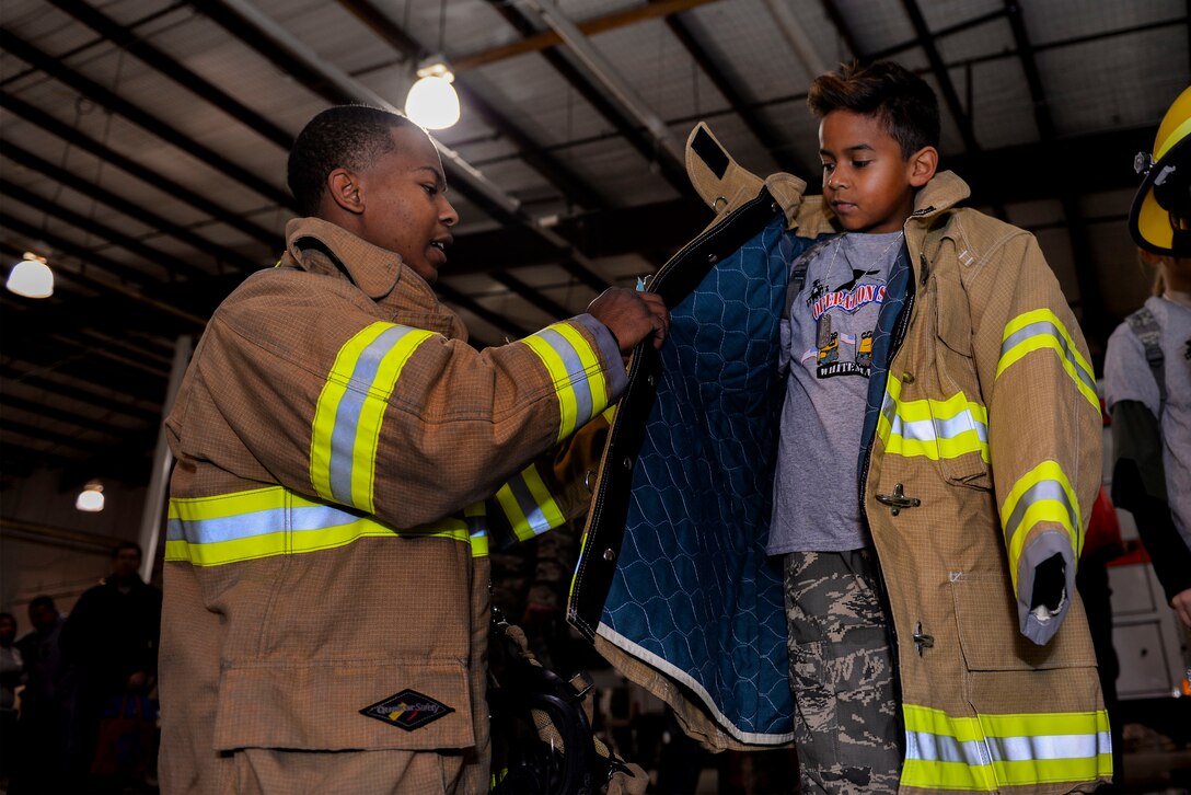 U.S. Air Force Airman 1st Class Christopher Walker, a 509th Civil Engineer Squadron firefighter, helps Nakai, the son of Tech. Sgt. David Brandon, the 509th Medical Operations Squadron mental health flight chief, try on protective gear during Operation Spirit at Whiteman Air Force Base, Mo., Nov. 21, 2015. Walker and other firefighters showed children how they help keep Airmen safe during deployments. (U.S. Air Force photo by Senior Airman Sandra Marrero)