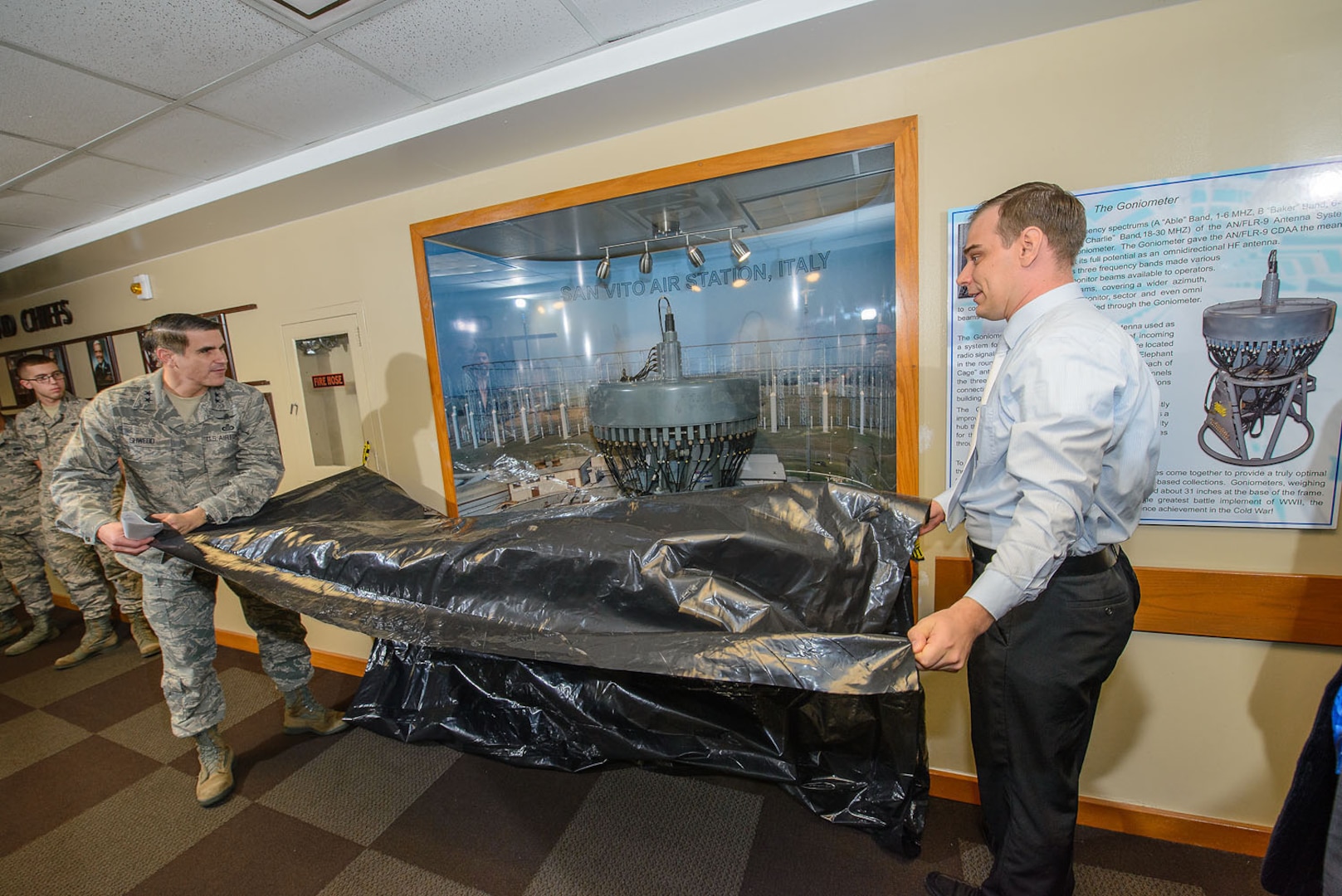 Maj. Gen. Bradford J. (BJ) Shwedo, 25th Air Force commander, and Mr. Justin Henderson, 25th A.F. directorate of history and research, pull off the plastic covering to unveil the new goniometer display, which is located in the main hallway of building 2000 at Joint Base San Antonio-Lackland, Texas. This goniometer was a part of the huge FLIR-9 antenna array at San Vito Air Station, Italy. (U.S. Air Force photo by William B. Belcher/Released)
