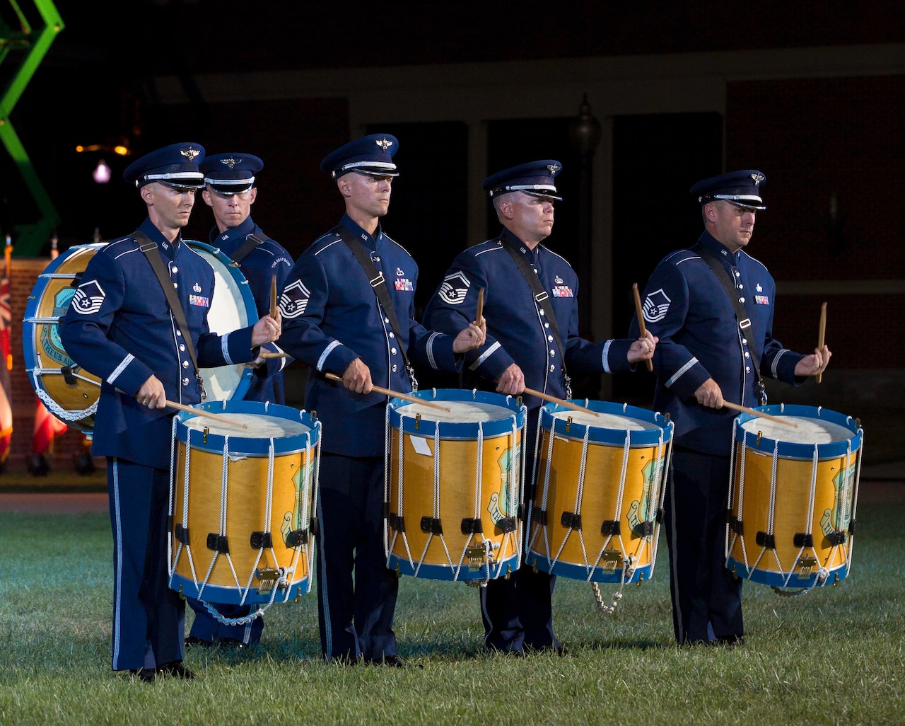 Four airmen in dress blue uniforms play large drums on a grassy field at night. A fifth airman walks past in the background.