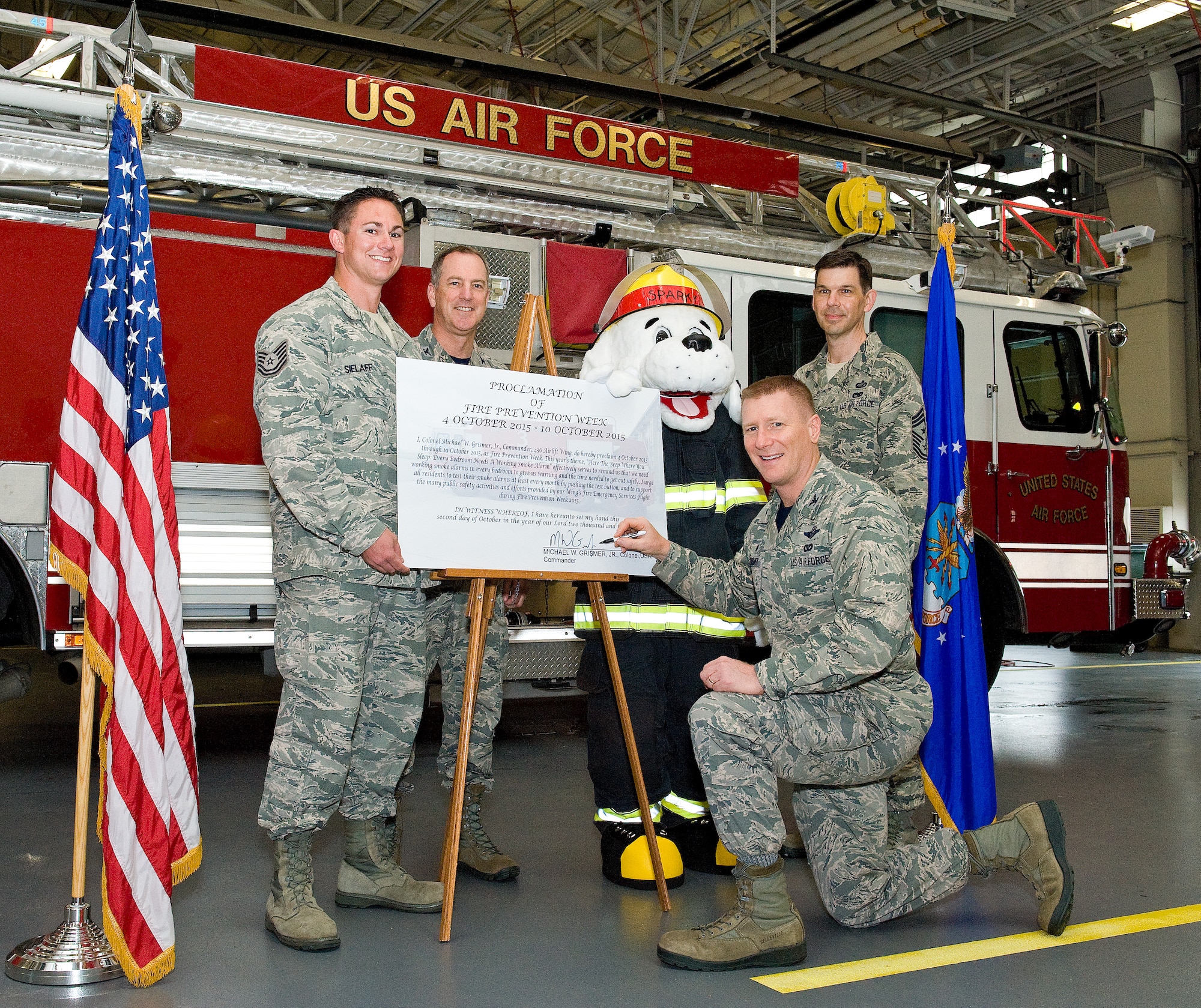 Col. Michael Grismer, 436th Airlift Wing commander, kneeling, signs the 2015 Fire Prevention Week proclamation Oct. 2, 2015, at the base fire department on Dover Air Force Base, Del. Standing left to right, Tech Sgt. Zachary Sielaff, 436th Civil Engineer Squadron fire inspector; Col. Robert Graham, 436th AW acting vice commander; Senior Airman Marc Winzeler, 436th CES fire department driver/operator as Sparky the fire dog; and Chief Master Sgt. Keith Davis, 436th AW command chief, witnessed the signing of the proclamation stating Oct. 4-10 as fire prevention week at Dover AFB and delivering a campaign theme of "Hear The Beep Where You Sleep: Every Bedroom Needs A Working Smoke Alarm." (U.S. Air Force photo/Roland Balik)