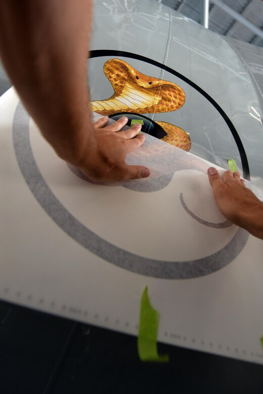 The carefully placed decal of a serpent coiled around a black sphere is slowly unveiled after application under the cockpit of an RC-135S Cobra Ball on Sept. 2, Offutt Air Force Base, Neb.  The design was created for the Cobra Ball by Duke Hemenway, graphic designer and illustrator from the Mississippi Gulf Coast area. (U.S. Air Force Photo by Josh Plueger/Released)