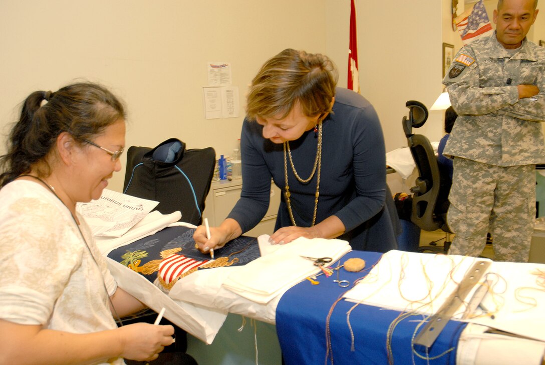 A DLA Troop Support Flag Room embroider lets DLA Foreign Policy Advisor Dolores Brown sign a presidential flag in-the-making, while DLA Senior Enlisted Leader Army Command Sgt. Maj. Charles Tobin looks on during a visit to the organization in Philadelphia Oct. 8, 2015. Both Tobin and Brown were impressed with the work DLA Troop Support employees do in support of the warfighter.