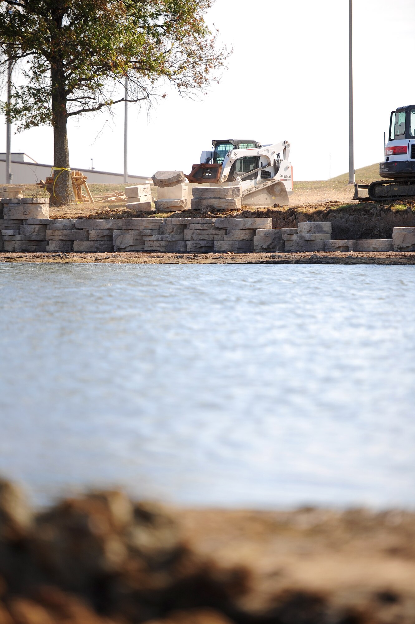 A contractor operates a Bobcat skid-steer loader to transfer bricks near the edge of Ike Skelton Lake Oct. 20, 2015, at Whiteman Air Force Base, Mo. To decrease algae and weed growth in shallow areas of the pond, the shoreline is being reinforced with a brick wall and is scheduled to be closed until December 2015. (U.S. Air Force photo by Airman 1st Class Jazmin Smith/Released)