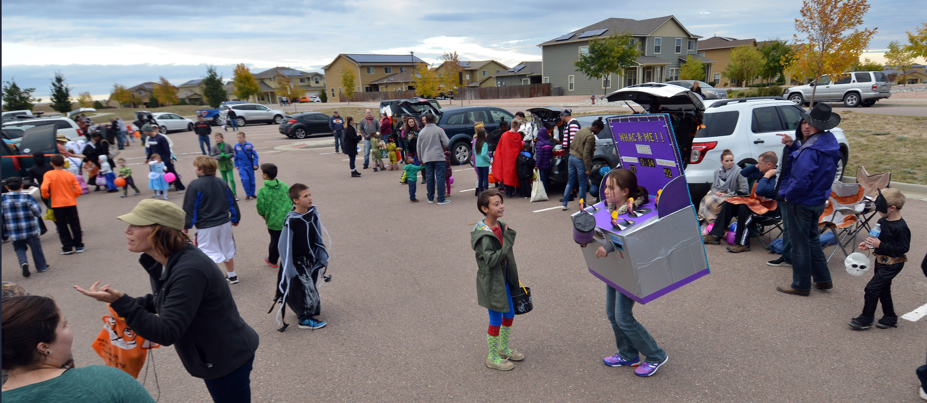 A crowd of parents and children dressed in Halloween outfits are trunk-or-treating. Meaning they walk around getting candy from cars that are parked in a wide circle (the photo takes place in a parking lot)