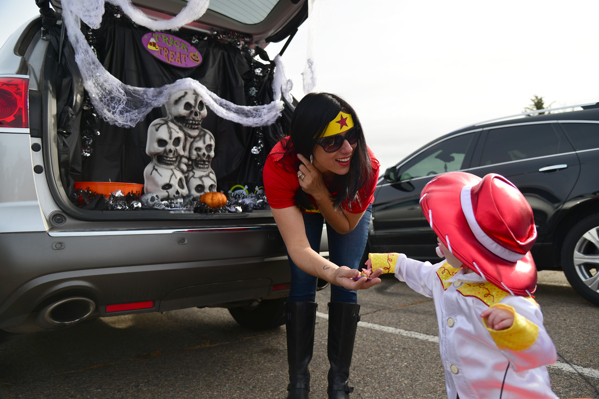 A woman hands a child a piece of candy at a trunk-or-treat. They are both dressed up in Halloween costumes. The woman as Wonder Woman and the little girl as Jessie from Toy Story.