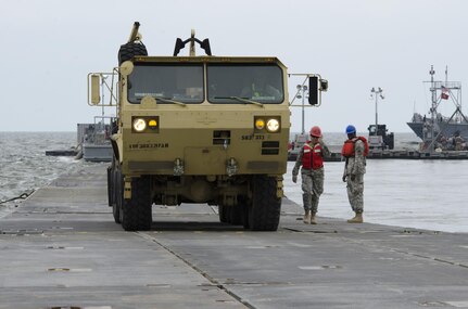 Cpl. Tauni Caldwell, 711th Transportation Company (Seaport Ops), acts as a walking guide for a heavy vehicle offloaded onto a floating causeway from a Landing Craft Mechanized watercraft (LCM) on Fort Story during Terminal Warrior 2015. The Logistics-Over-the-Shore (LOTS) exercise took place in the Hampton Roads area of Virginia from mid July through mid August and included Reserve, National Guard and Active Duty units focused on cohesion and sharpening proficiency in basic LOTS skills as part of the Total Army Concept