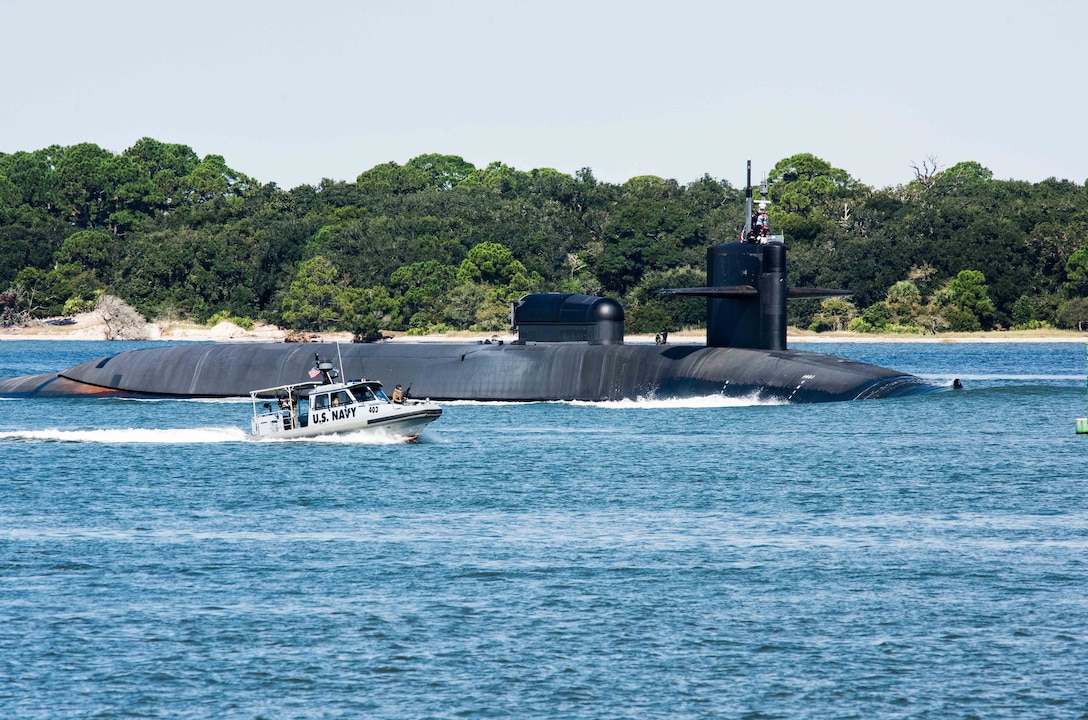 A large submarine, partially submerged, sails through the ocean escorted by a small boat that reads “U.S. Navy” on its side. An uninhabited beachfront is in the background.