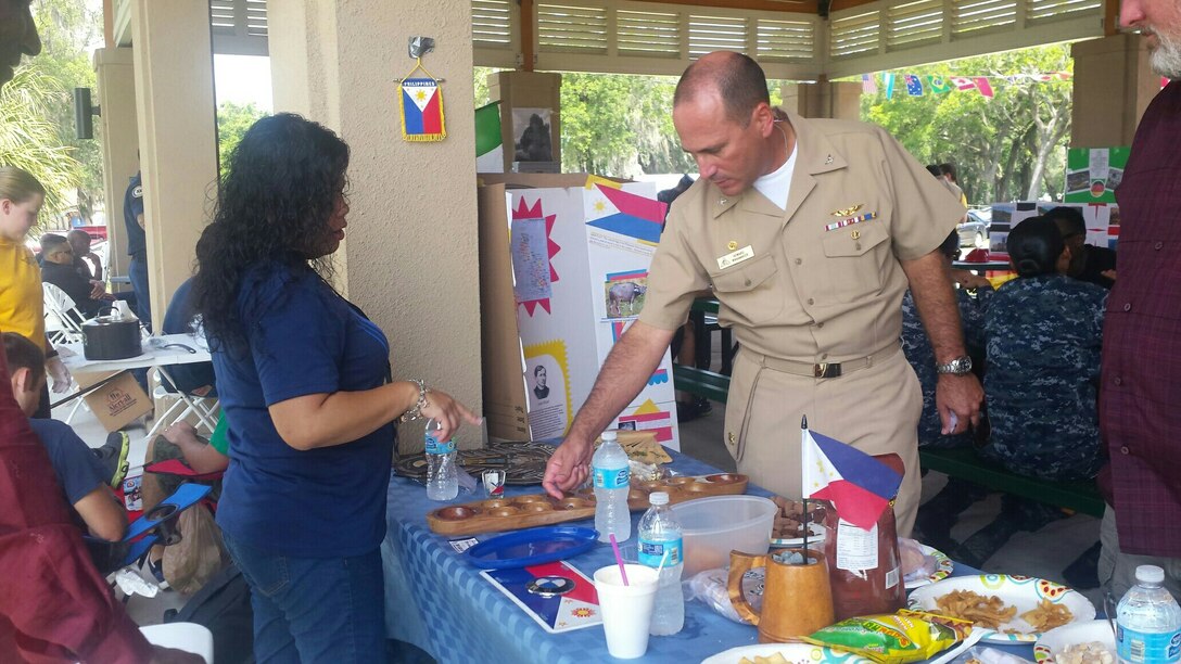 Naval Air Station Jacksonville, Florida Commander Captain Howard Wanamaker visits Defense Logistics Agency Aviation's display on the Philippines during the base’s Multicultural Fun Day July 22, 2015. DLA Aviation at Jacksonville Sustainment Specialist Leslie Stanley provides the commander with instructions on how to play Sungka, one of the most popular board games in the Philippines.