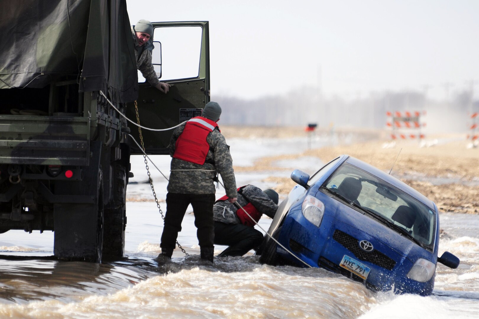 Sgt. Preston Steele, right, and Spc. Jeremy Kasperson, both of the 815th Engineer Company, attach a towing chain to a partially submerged vehicle March 25 on a washed out portion of a gravel road a few miles west of Harwood, N.D., Cass County Tactical Operations Center liaison officer Capt. Grant Larson, of the 119th Maintenance Squadron, North Dakota Air National Guard, can be seen giving guidance from the cab of the truck. Steele and Kasperson are members of a North Dakota National Guard quick response force team that is being called to the scene by the Cass County Sherriff's Department to assist with the vehicle recovery. The driver of the vehicle was able to get clear of the car and make his way through the freezing water to safety before help arrived. (North Dakota Air National Guard photo by Senior Master Sgt. David Lipp)