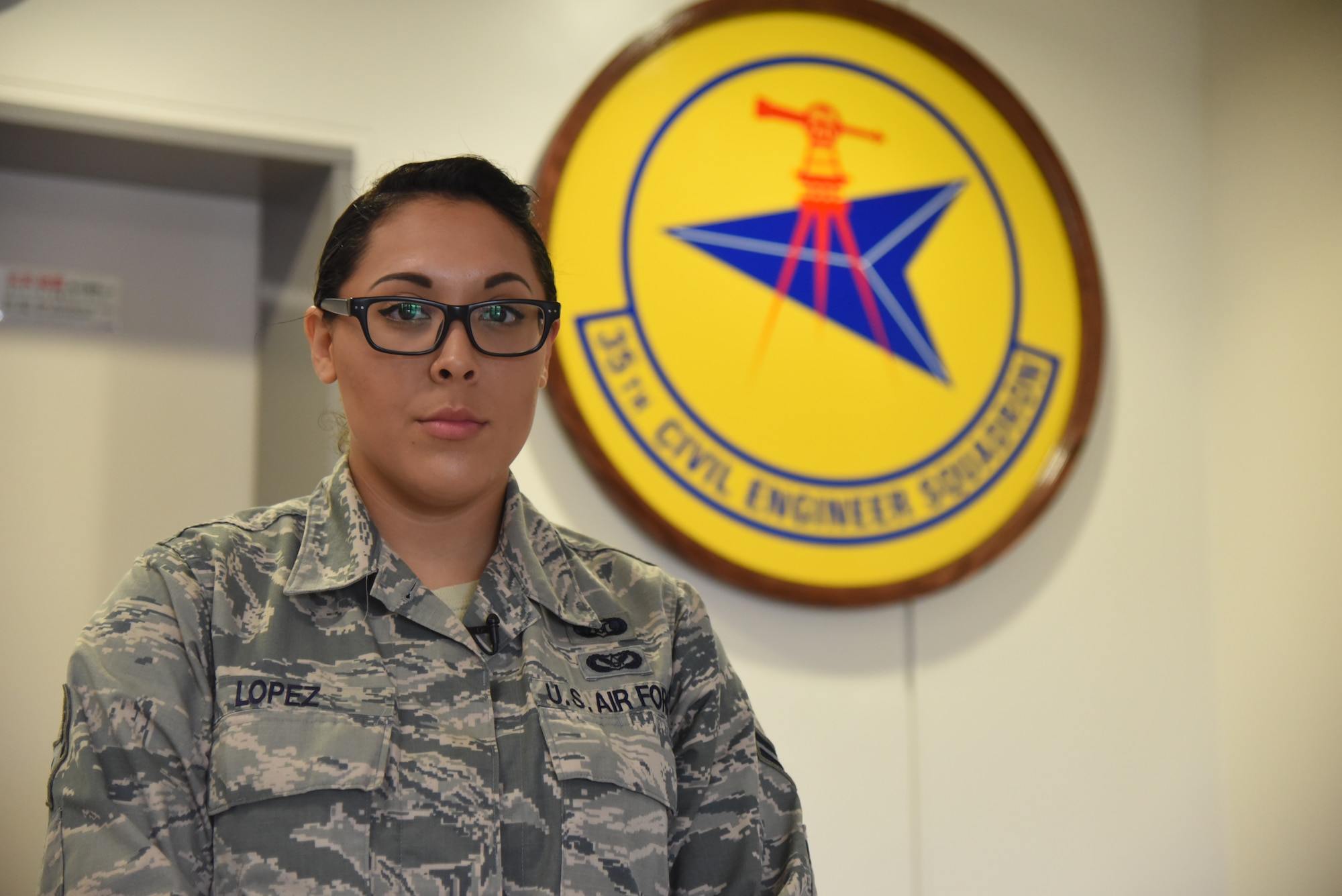 U.S. Air Force Airman 1st Class Joanna Lopez, 35th Civil Engineer Squadron emergency management logistics journeyman, poses in front of the 35 CES emblem at Misawa Air Base, Japan, Sept. 10, 2015. As an emergency management logistics journeyman, Lopez ensures Airmen are prepared for any potential disaster situations by instructing courses on safety and is an important component during 35th Fighter Wing exercises, executing safety procedures are followed without discrepancy. (U.S. Air Force photo by Senior Airman Patrick S. Ciccarone/Released)