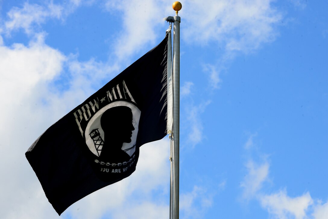 The Prisoner of War/Missing in Action flag waves during the Joint Base Langley-Eustis, Va., POW/MIA Recognition Day Ceremony at Langley Air Force Base, Va., Sept. 18, 2015. The black and white image of a gaunt silhouette, a strand of barbed wire and an ominous watchtower, on the flag was designed by Newt Heisley, a former World War II pilot. (U.S. Air Force photo by Senior Airman Kimberly Nagle/Released) )