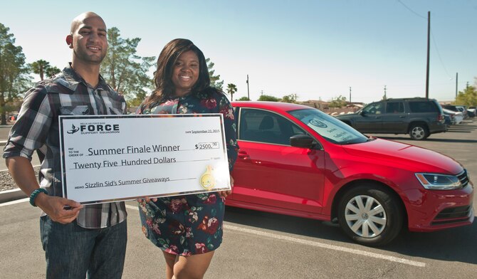 Carlos Del Valle, 99th Force Support Squadron Outdoor Recreation Center outdoor adventure programmer, poses with a $2,500 check presented to him by Takeira Jones-Bolden, 99th FSS supervisory marketing specialist and commercial sponsorship coordinator, in front of the brand new 2015 Volkswagen Jetta he won at Nellis Air Force Base, Nev., Sept. 23, 2015. Del Valle won the 99th FSS’ Ultimate Summer Giveaway grand prize of the new car by having his name drawn during the car’s raffle. Del Valle was one of almost 10,000 Nellis and Creech AFB members who entered the drawing, which began in June. In addition to winning the car, Del Valle also was given $2,500 spending cash to pay for the car’s tax, title and licensing fees. (U.S. Air Force photo by Staff Sgt. Siuta B. Ika)