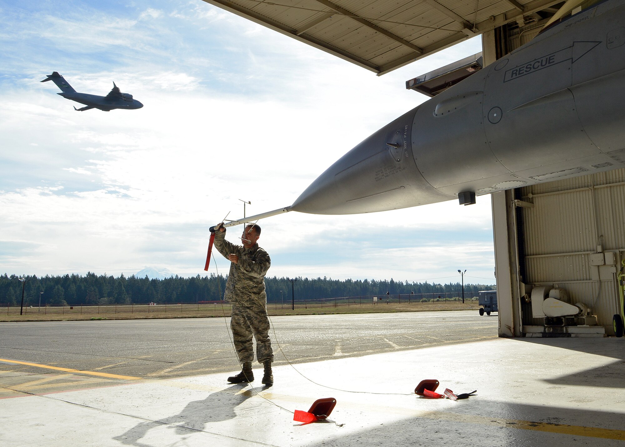 As a C-17 Globemaster III passes by in the background, Senior Airman Andres Elias (left), 412th Aircraft Maintenance Squadron assistant dedicated crew chief, covers the test instrumentation pitot tube of an Edwards Air Force Base F-16 Falcon on Sept. 24, 2015 at Joint Base Lewis-McChord, Wash., in preparation for the KC-46 Pegasus first flight. Elias is just one of 20 Edwards AFB personnel temporarily assigned to JBLM in support of the KC-46 Tanker Program. (U.S. Air Force photo by Jet Fabara)