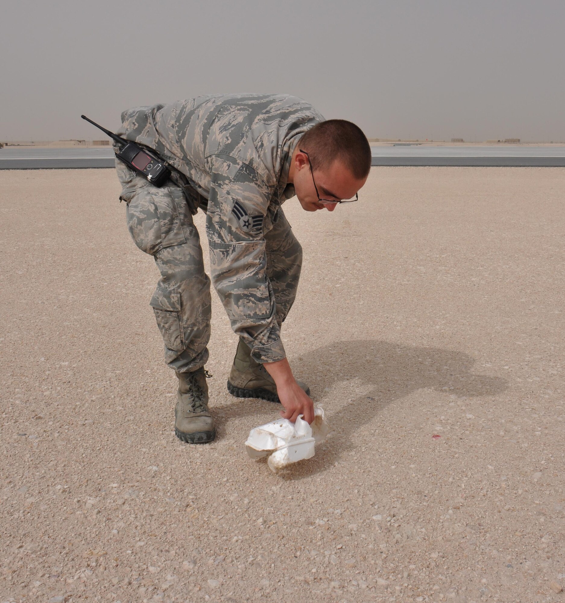 Senior Airman Logan Winter, 379th Expeditionary Operations Support Squadron Airfield Management journeyman from Quincy, Washington, picks up a piece of trash near a runway at Al Udeid Air Base, Qatar March 28. Winter conducts several checks of the airfield each day to ensure foreign objects and debris are removed. In 2015, the 379 EOSS Airfield Management team supported more than 20,000 sorties. Photo altered for security purposes. (U.S. Air Force photo by Tech. Sgt. James Hodgman/Released)