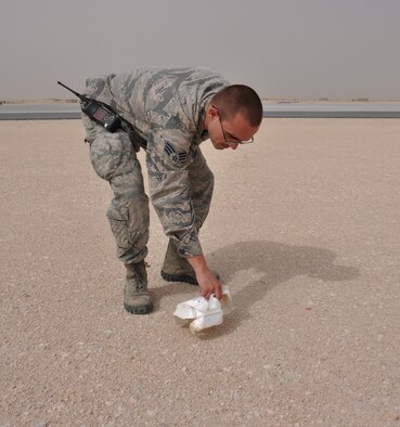 Senior Airman Logan Winter, 379th Expeditionary Operations Support Squadron Airfield Management journeyman from Quincy, Washington, picks up a piece of trash near a runway at Al Udeid Air Base, Qatar March 28. Winter conducts several checks of the airfield each day to ensure foreign objects and debris are removed. In 2015, the 379 EOSS Airfield Management team supported more than 20,000 sorties. Photo altered for security purposes. (U.S. Air Force photo by Tech. Sgt. James Hodgman/Released)