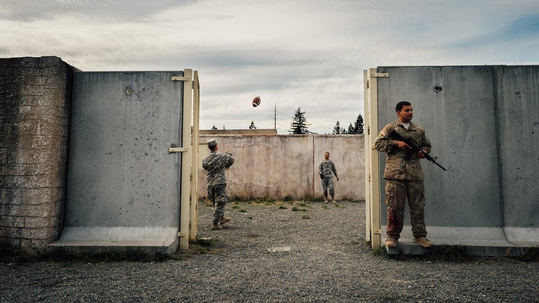 Sports Photography: Two soldiers play catch with a football while a fellow soldier watches the perimeter of the training grounds during the Expert Field Medic Badge course at Joint Base Lewis-McChord, Wash., Sept. 24, 2015. The EFMB is the non-combat equivalent of the Combat Medical Badge and is awarded to medical personnel of the U.S. military who successfully complete a set of qualification tests. U.S. Air Force photo by by Senior Airman Jordan A. Castelan