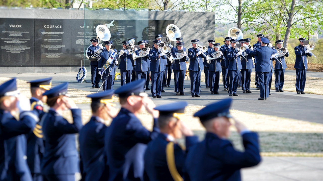Band members dressed in military blue uniforms play horns, tubas and other instruments in front of the U.S. Air Force Memorial as other airmen salute.