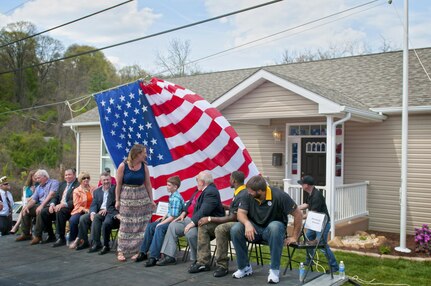 Staff Sgt. Michelle Satterfield, with the 14th Quartermaster Detachment, stands up in surprise as her new house is revealed behind the American flag during her ceremony in McKeesport, Pa., April 25, 2016. Satterfield is receiving a house built by the students of the McKeesport Area High School and Stephen Siller Tunnel to Towers Foundation for all of the volunteer work she has done in the past years. (U.S. Army photo by Staff Sgt. Dalton Smith / Released)