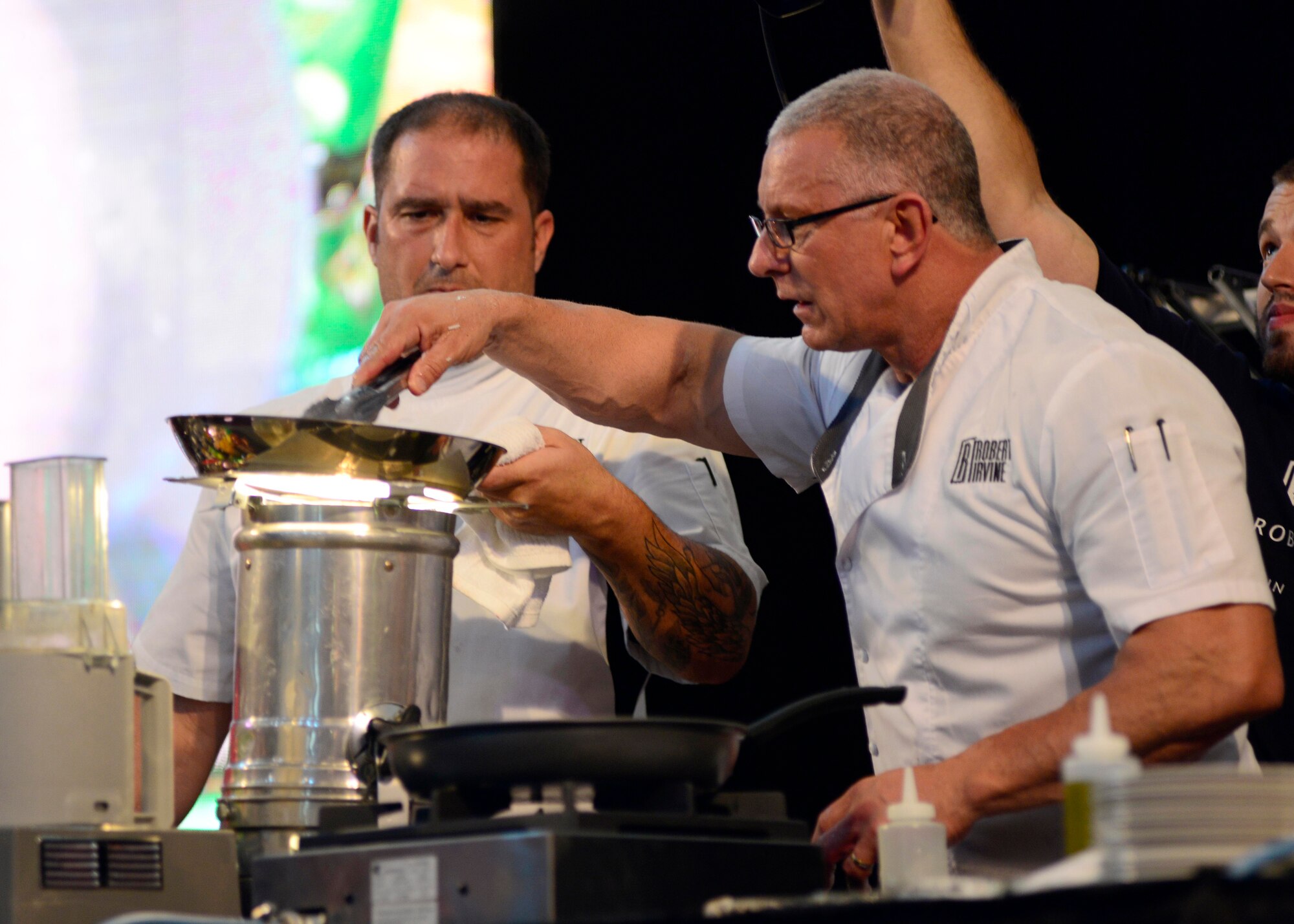 Robert Irvine (right), celebrity chef, uses a stage light to cook steak, during a live show, July 30, 2016 at Aviano Air Base, Italy. Irvine visited Team Aviano as part of a live USO Tour to thank service members and their families for their service. (U.S. Air Force photo by Senior Airman Areca T. Bell/Released)