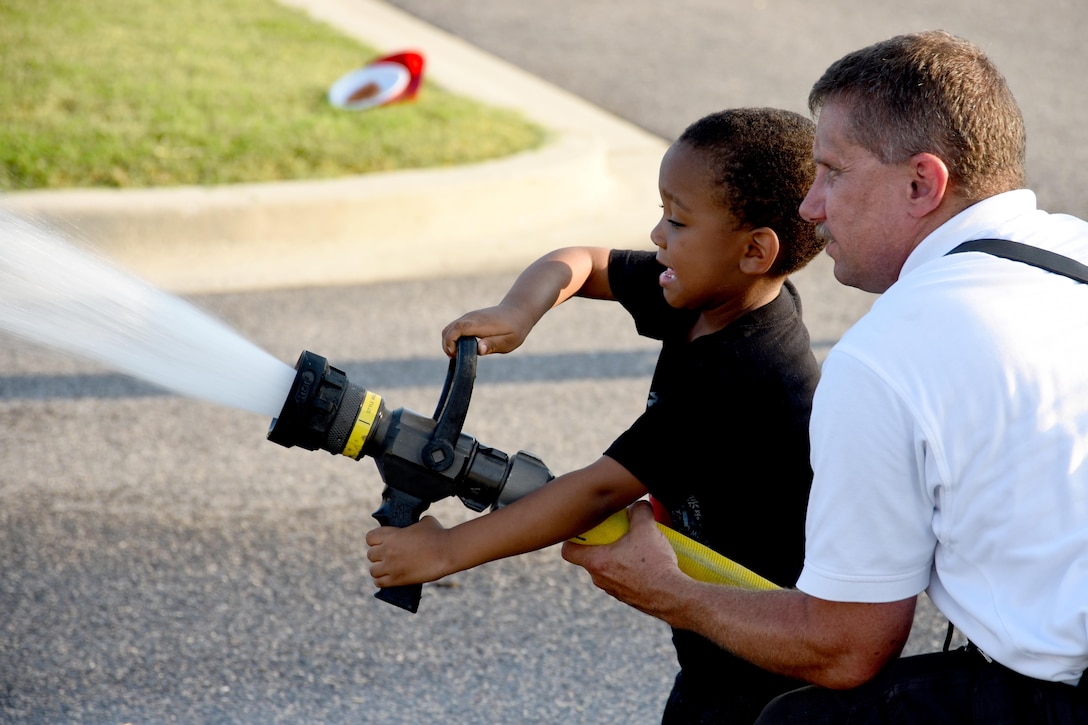 Roy McKlveen, Joint Base Andrews Fire Department Station one station captain and volunteer firefighter with the Morningside Volunteer Fire Department, assists Gabriel Cole, grandson of retired Tech. Sgt. Tara Mundy, with a fire hose during a National Night Out event held at the Liberty Park welcome center on JBA, Md., Aug. 2, 2016. NNO was established in 1984 and developed as a crime prevention program that emphasizes building partnerships between the police and the community. (U.S. Air Force photo by Airman Gabrielle Spalding)