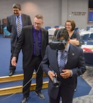 William Parks, a contractor supporting the Virtual Reality training team, assists Richard Perez, President, San Antonio Chamber of Commerce, with virtual reality goggles as Doctor Ricardo Romo, President, University of Texas, San Antonio, looks on.
