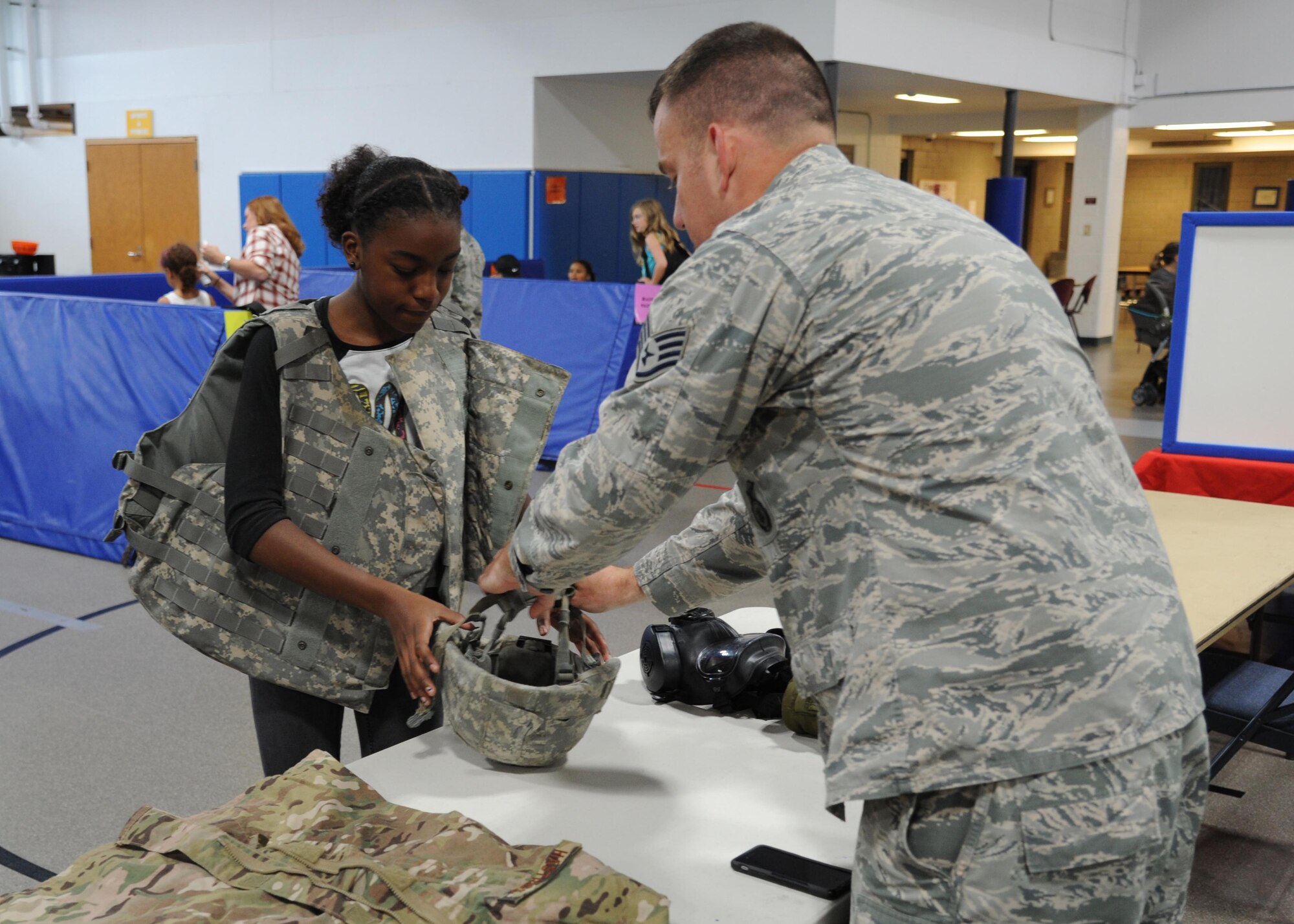 Camille Edwards, 12, left, tries on personal protective equipment with the assistance of Staff Sgt. James Hastings, 319th Security Forces Squadron combat arms instructor, right, August 4, 2016, at Liberty Square on Grand Forks Air Force Base, N.D. Hastings was one of many volunteers who provided entertainment and education at the School Liaison Office Back to School Picnic. (U.S. Air Force photo by Senior Airman Ryan Sparks/Released)