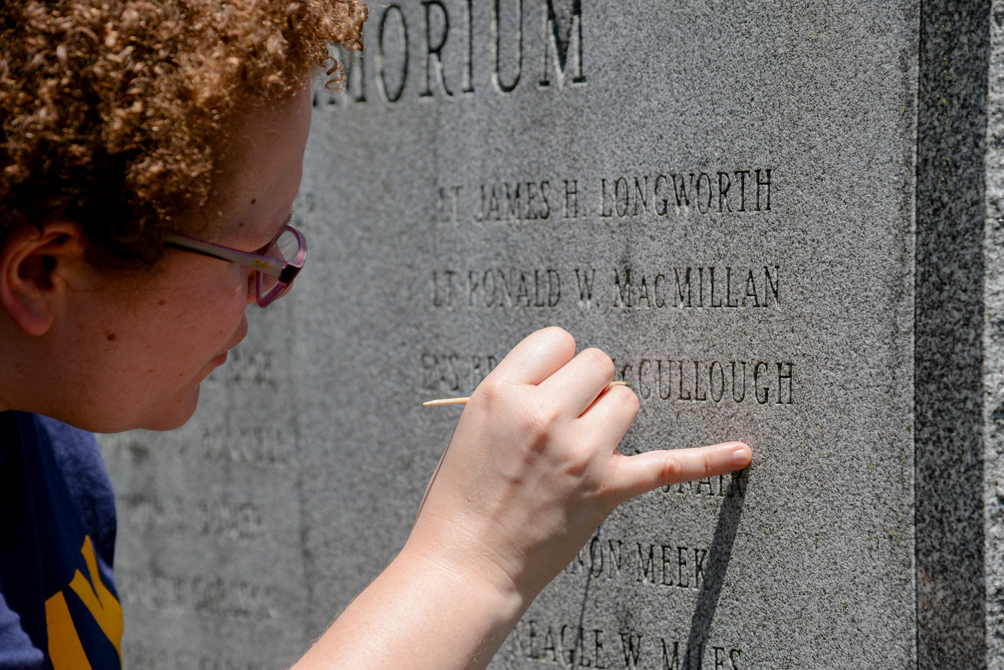 A picture of Melissa Swanson, Naval History and Heritage Command Conservator, using a wooden pick to remove lichens growing inside the letters of inscriptions on a U.S. Navy VC-4 memorial marker.
