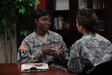 A female Social Worker wearing ACU sitting inside at table and talking with female Soldier wearing ACU.
