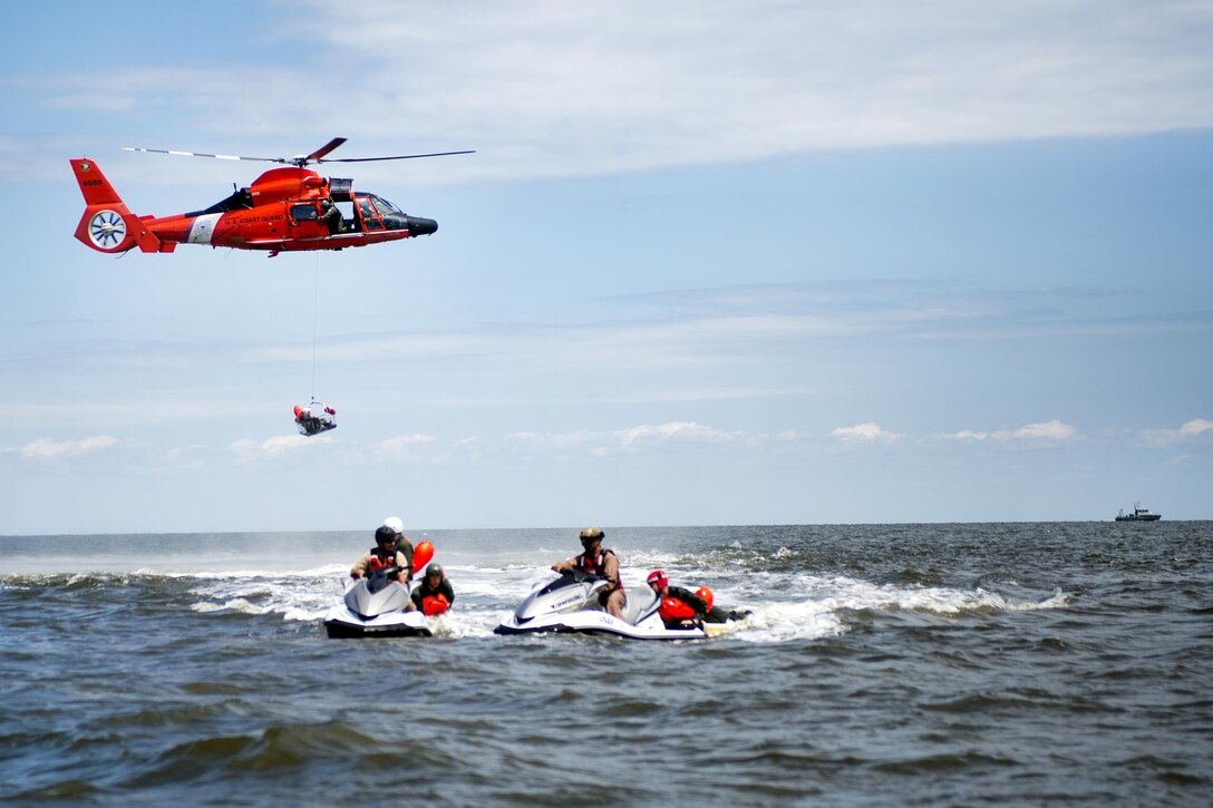 Water scooter operators transport airmen on the back of their vehicles while a Coast Guard H-65 Dolphin helicopter hoists another airman in a basket during water survival training at Bowers Beach, Del., Aug. 6, 2016. Air Force photo by Tech. Sgt. Nathan Rivard