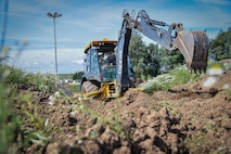 Chief Master Sgt. Edwin Ludwigsen, 52nd Fighter Wing command chief, uses a backhoe to dig holes under the guidance of Airman 1st Class Justin Leyno, 52nd Civil Engineer Squadron heavy equipment operator, during an “out and about” visit at Spangdahlem Air Base, Germany, Aug. 11, 2016. (U.S. Air Force photo by Tech. Sgt. Christopher Parr)