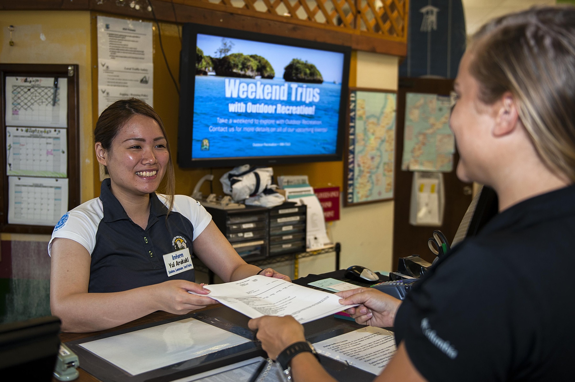 Yui Arakaki, 18th Force Support Squadron intern, hands an informational packet to a customer Aug. 24, 2016, at Kadena Air Base, Japan. KLI interns have the opportunity to build ties between Kadena and the community while practicing their English skills. (U.S. Air Force photo by Airman 1st Class Corey M. Pettis)