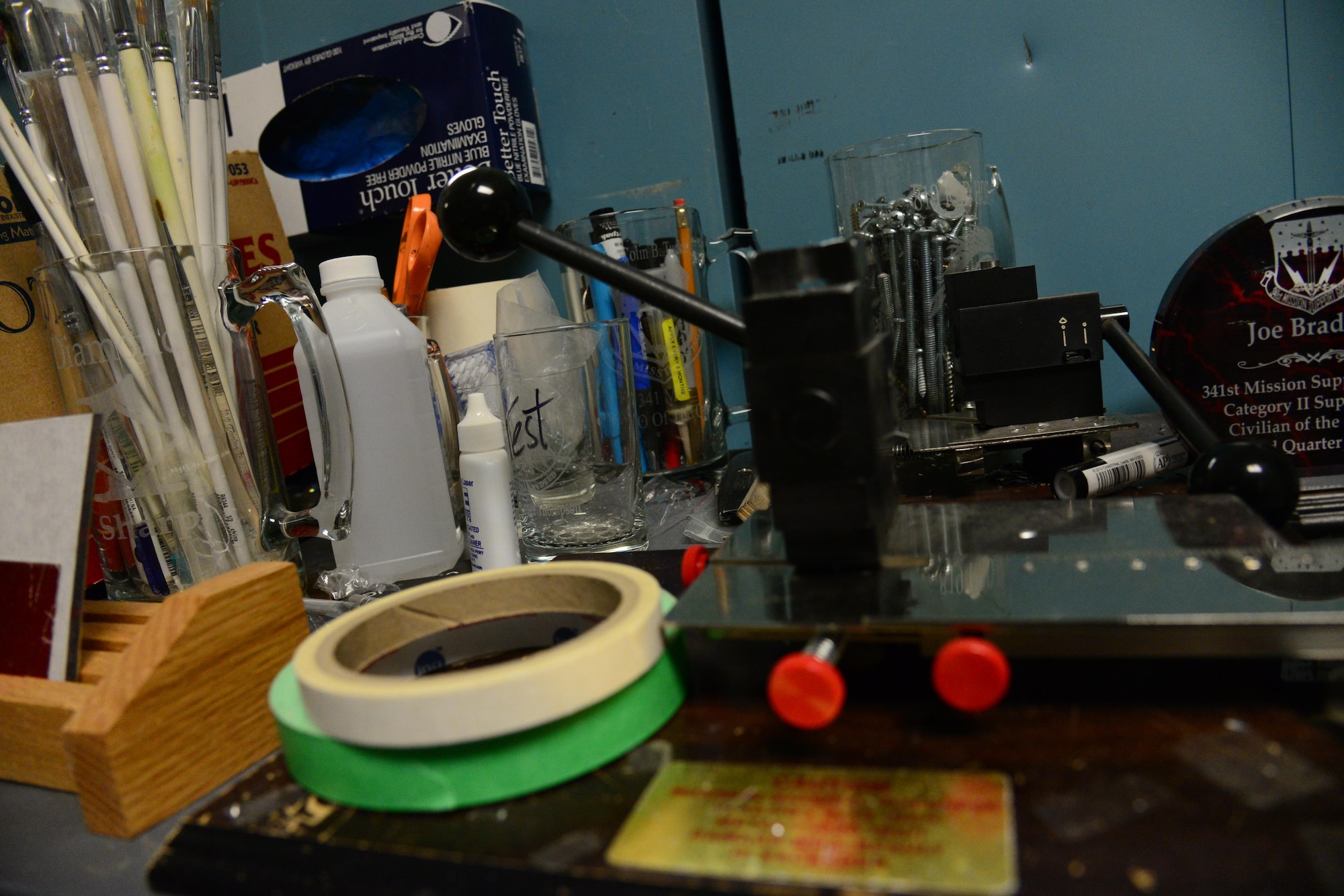 A crowded work desk the engraves uses located at the Engraving Shop in the Arts and Crafts Center at Malmstrom Air Force Base, Mont., Dec. 7, 2016. The Engraving Shop uses a plethora of products and tools to enable them to make most of their plaques, trophies and awards from scratch. (U.S. Air Force photo/Airman 1st Class Magen M. Reeves)