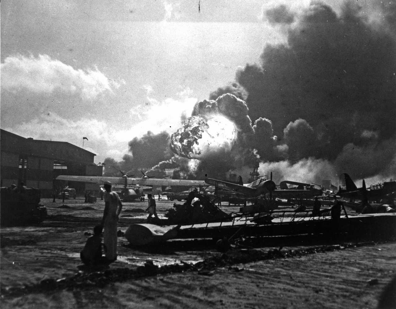 Sailors at Ford Island Naval Air Station, Hawaii, look on as the USS Shaw explodes in the distance, Dec. 7, 1941. This view is of the PBY5-A Catalina ramp with assorted aircraft scattered among the debris. Barely seen in the background is the beached USS Nevada. National Park Service photo