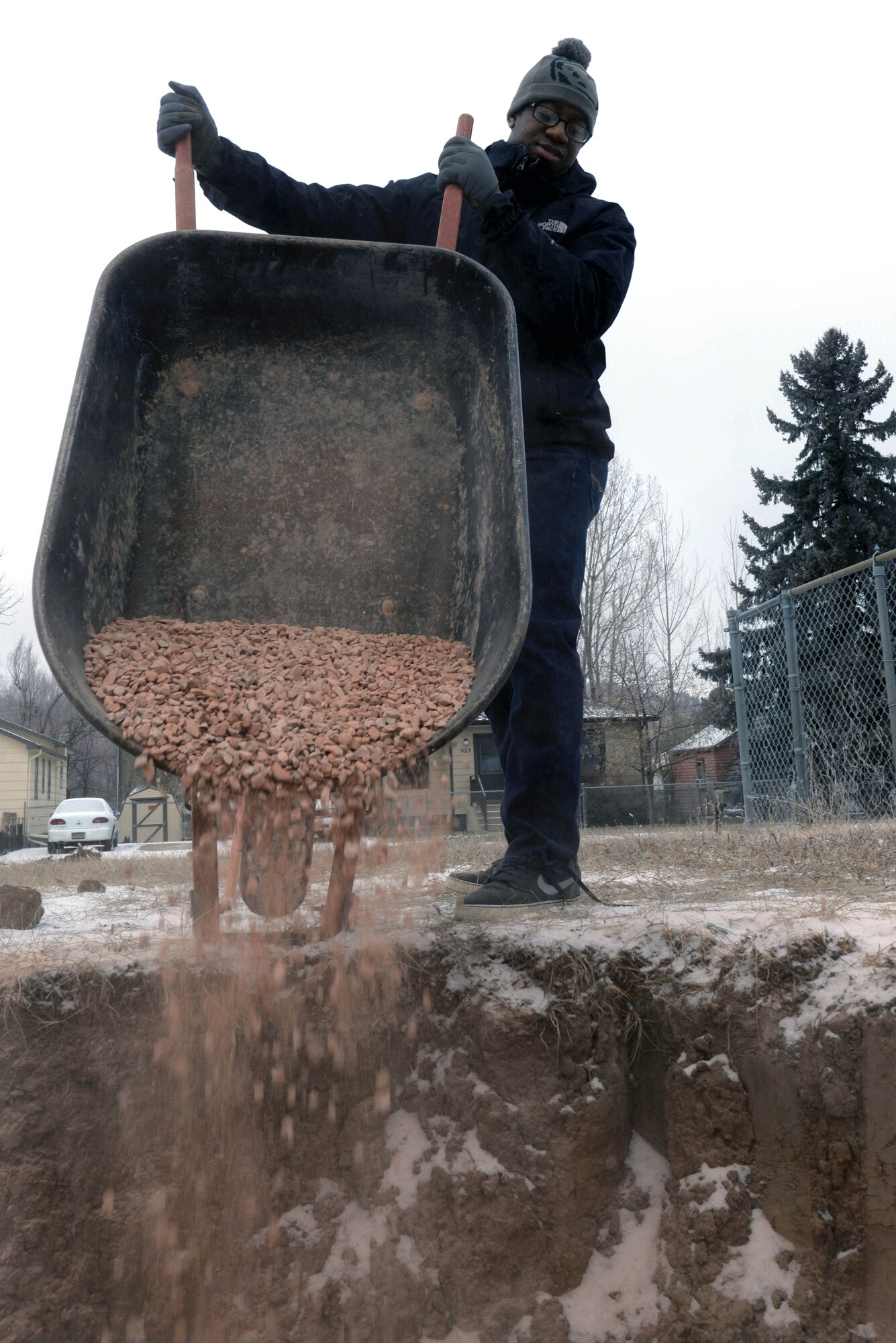 Senior Airman Shaquille Robinson, an intel analyst assigned to the 89th Attack Squadron, pours gravel next to a housing foundation to support its piping in Rapid City, S.D., Dec. 10, 2016. Piping installed in uncompacted ground can develop negative pitch causing stress on the pipe, laying the pipe on a gravel bed will resolve the issue. (U.S. Air Force photo by Airman 1st Class Donald C. Knechtel)