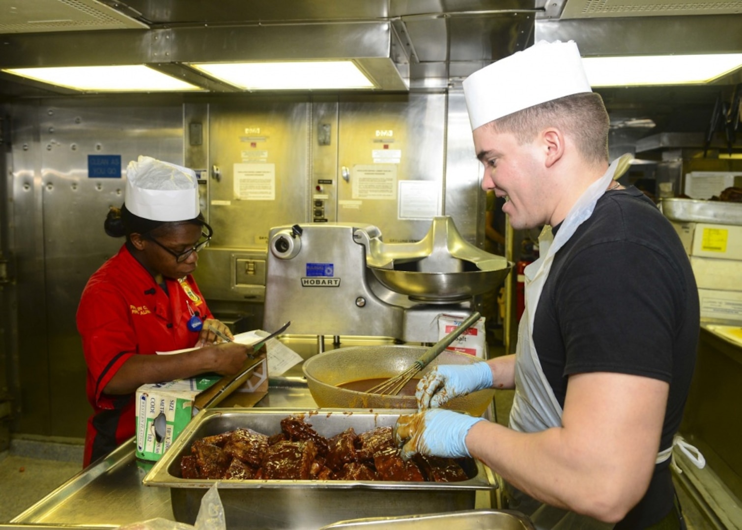 Marine Corps Cpl. Giovanni Perez, right, prepares barbecued ribs in the galley aboard the amphibious dock landing ship USS Whidbey Island, Nov. 14, 2016. Navy photo by Petty Officer 2nd Class Nathan R. McDonald