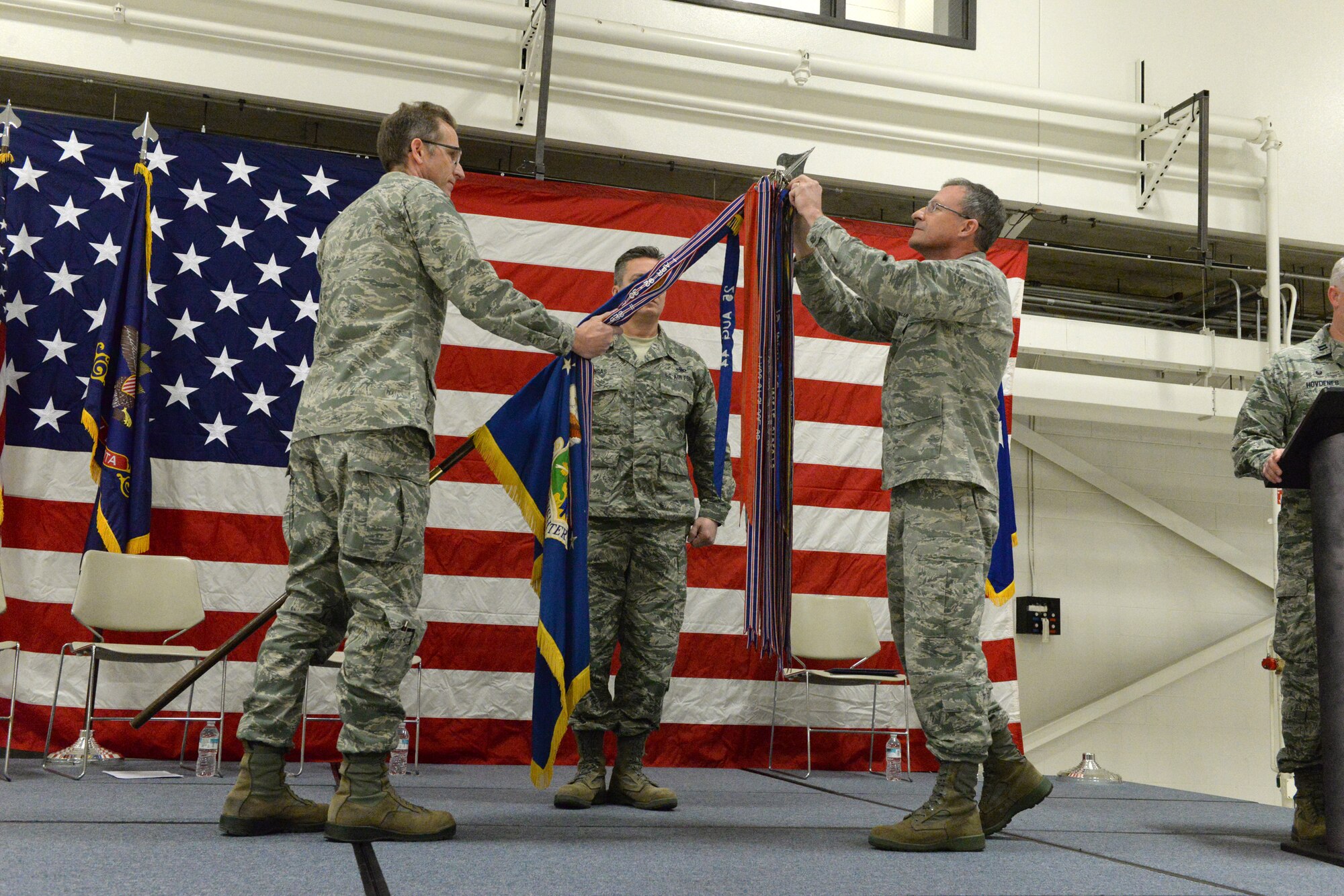 Brig. Gen. Robert Becklund, the North Dakota deputy adjutant general, right, attaches a streamer, representing the Air Force Outstanding Unit Award, onto the 119th Wing guidon, as Col. Kent Olson, the 119th Wing commander, lowers the unit flag during a recognition ceremony at the as Chief Master Sgt. Duane Kangas, the 119th Wing command chief master sergeant, center, looks on at the North Dakota Air National Guard Base, Fargo, North Dakota, Dec. 3, 2016. (U.S. Air National Guard photo by Senior Master Sgt. David H. Lipp/Released)